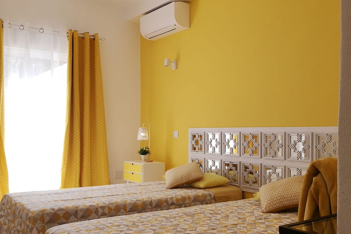 A hotel room with two beds, a yellow wall, a patterned headboard, and a hanging light fixture.