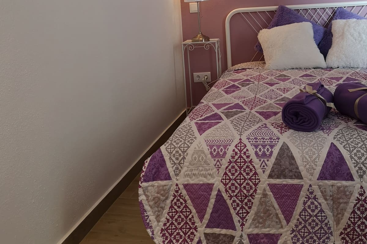 A bedroom with a bed, a nightstand, a lamp, and a wall-mounted shelf with decorative items.