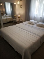 Una cama bien hecha con un espejo grande arriba, dos lámparas de noche y una mesita redonda con una lámpara.