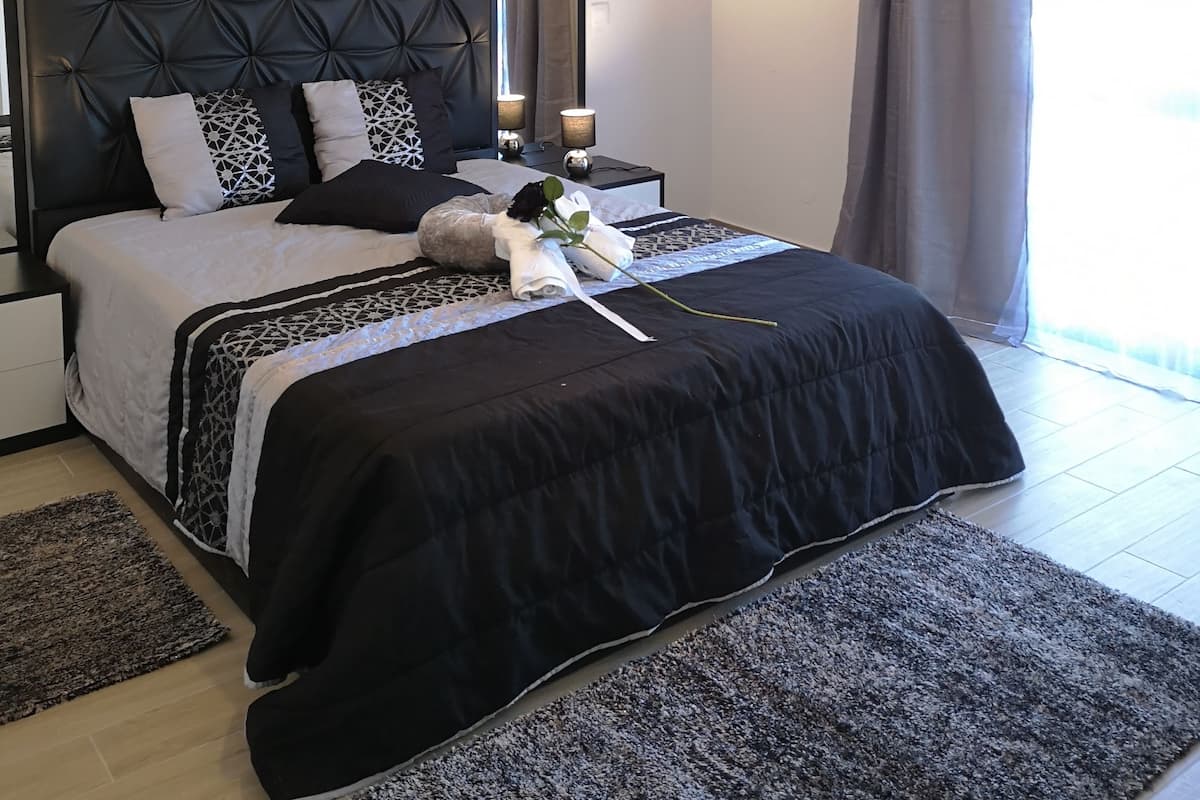 A modern bedroom with a tufted headboard, a large bed with a black and white bedspread, two bedside tables with lamps, and a large window with curtains.