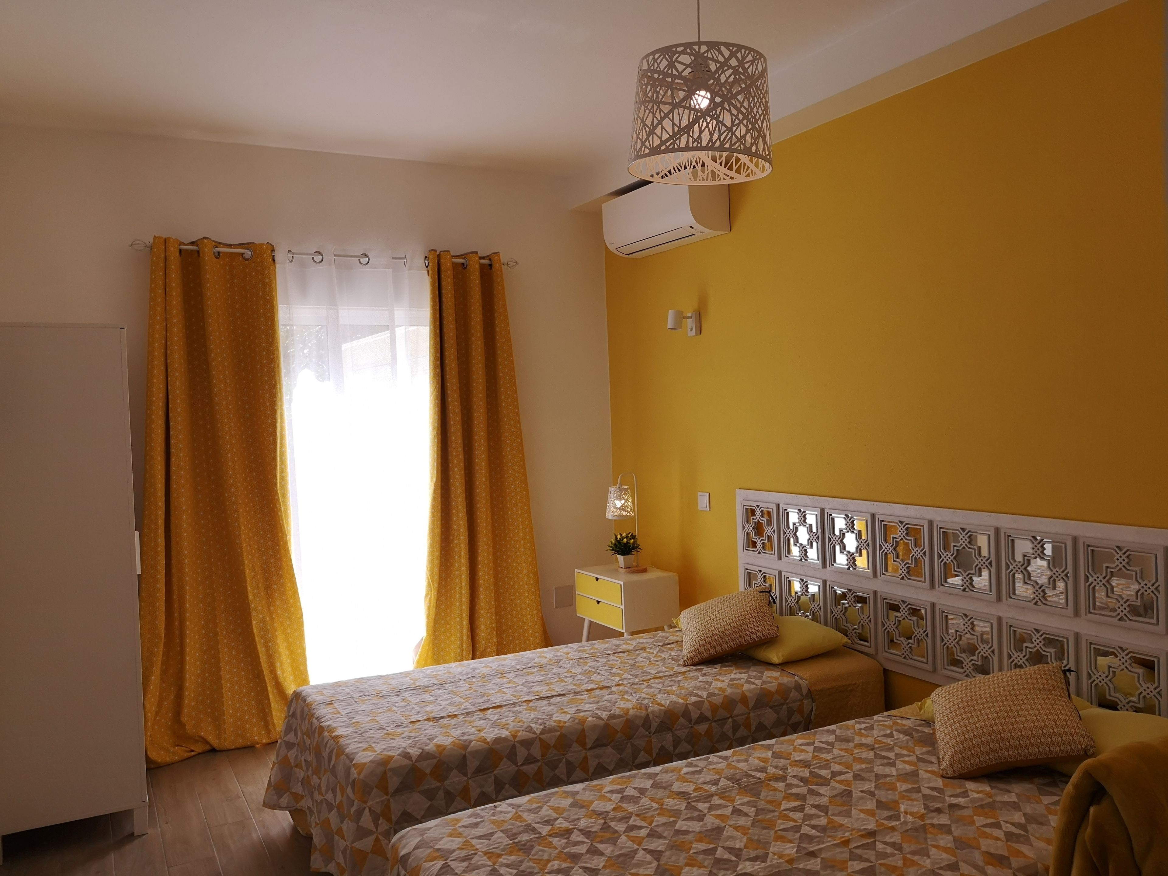 A bedroom with two beds, a window with curtains, an air conditioning unit, a bedside table with a lamp, and decorative wall panels.
