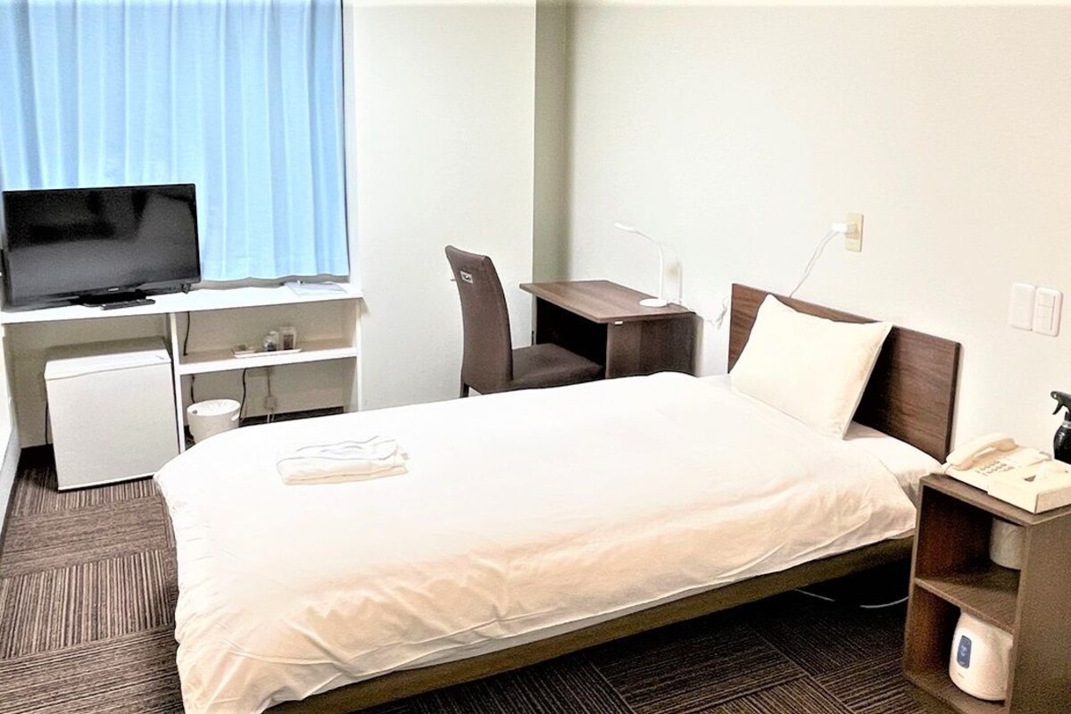 A hotel room with a bed, desk, chair, television, and a small kitchenette.