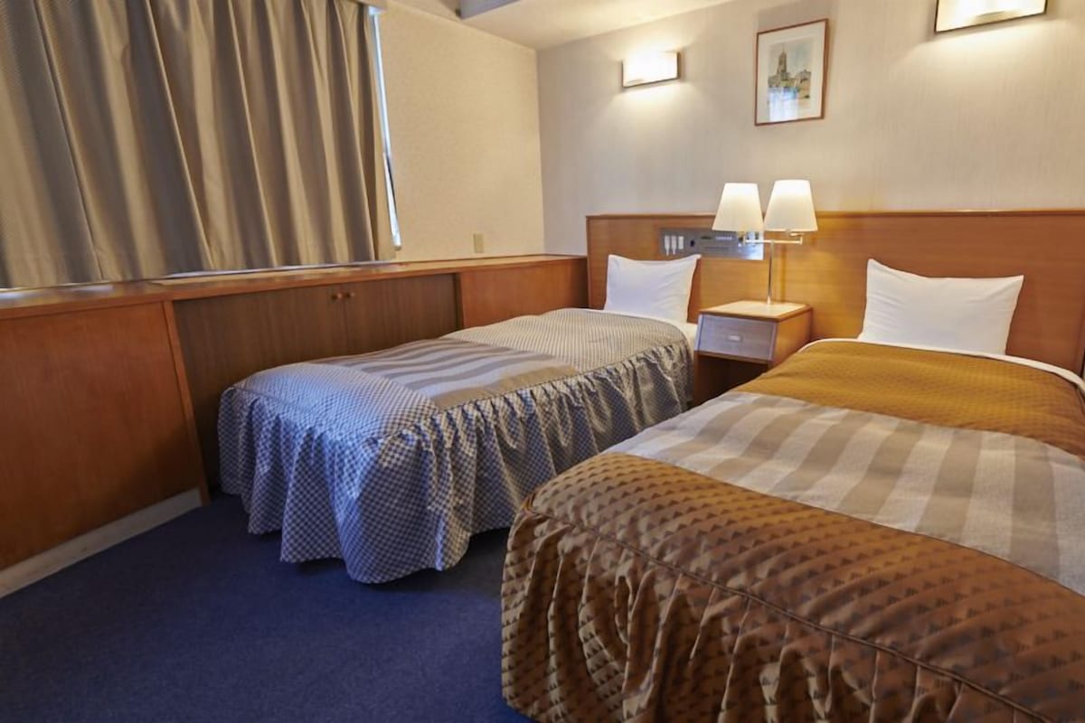 A hotel room with two single beds, a wooden headboard, a nightstand with lamps, and a window with curtains.