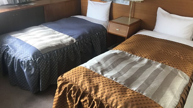 A hotel room with two single beds, each with a patterned bedspread, a wooden headboard, bedside tables with lamps, and a nightstand with a lamp. - GOI Park Hotel (Ichihara)