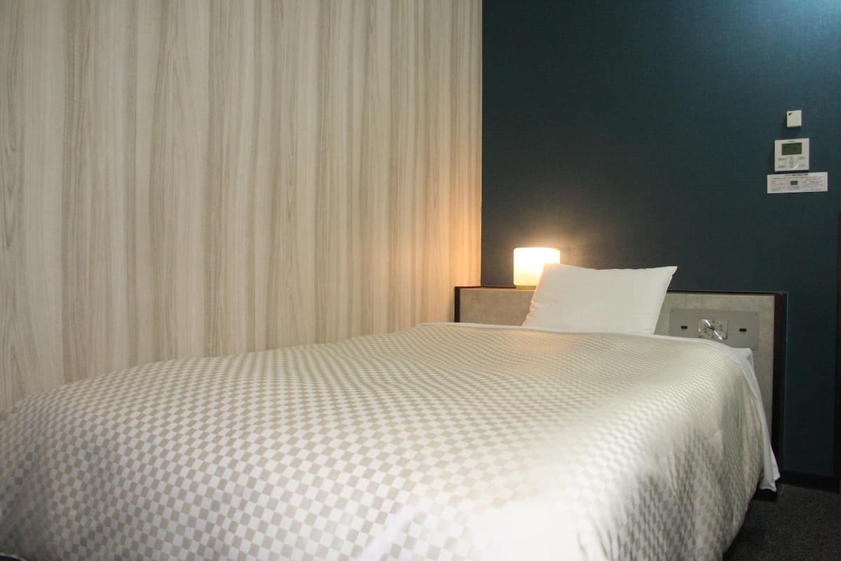 A hotel room with a bed, a nightstand, a lamp, and a wall-mounted thermostat.