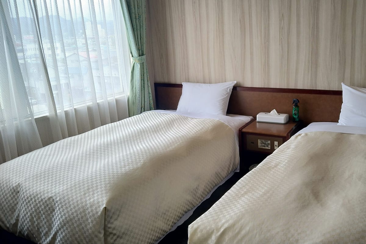 A hotel room with two beds, a nightstand, and a window with sheer curtains.