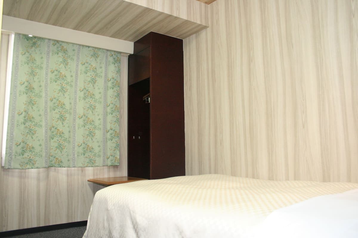 A hotel room with a bed, a wooden nightstand, a wardrobe, and a window with floral curtains.
