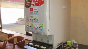 A Coca-Cola vending machine with multiple drink options, a sign indicating 'Welcome Drink 15:00~21:00', and a chair in the foreground.