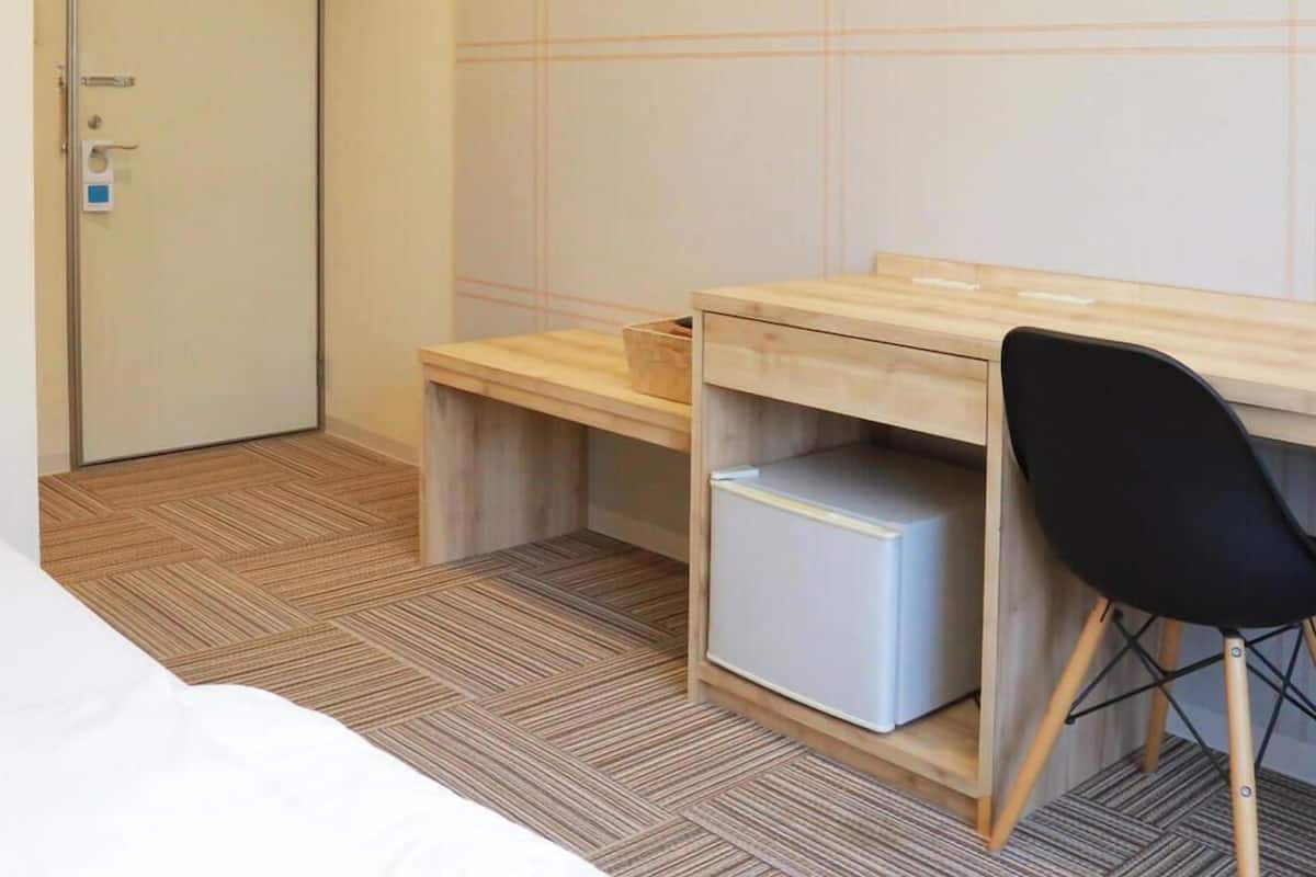 Ein Hotelzimmer mit Bett, Schreibtisch mit Stuhl, kleinem Kühlschrank und einer Tür.