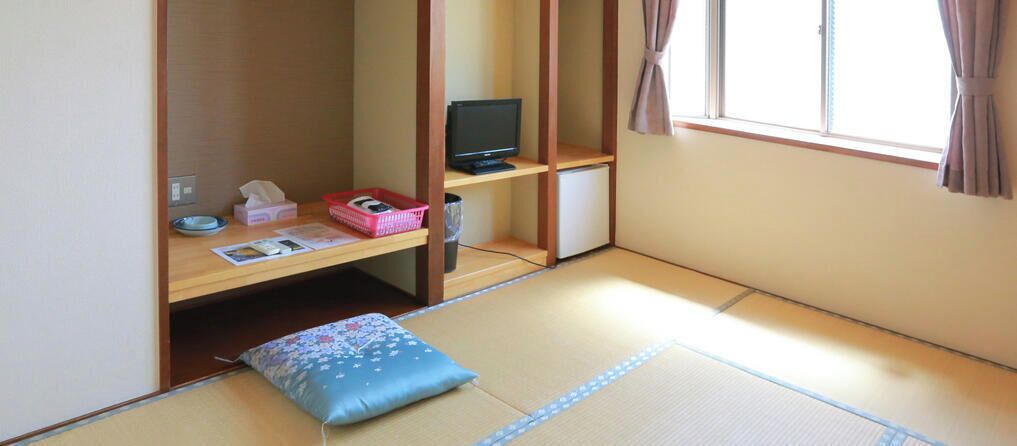 A tatami mat room with a futon bed, a television, a small desk with a computer, and a window with curtains.