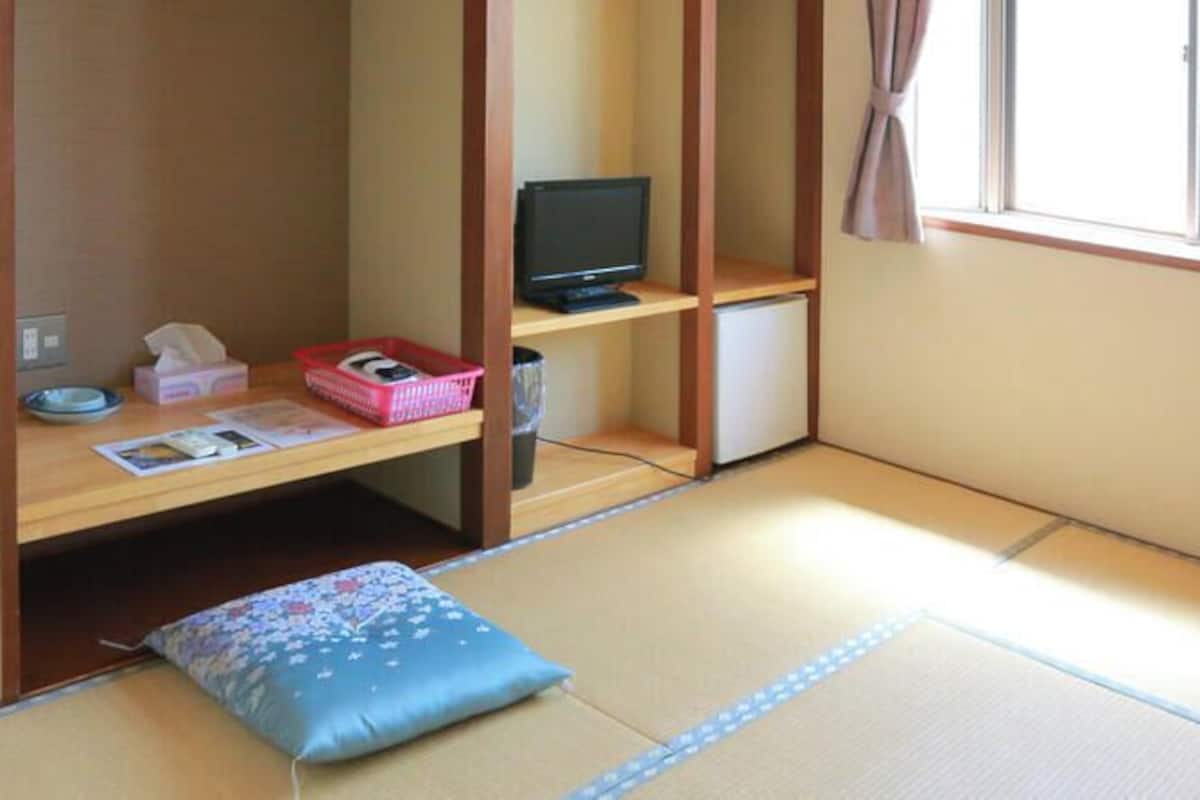 A tatami mat room with a futon bed, a television, a small desk with a computer, and a window with curtains.