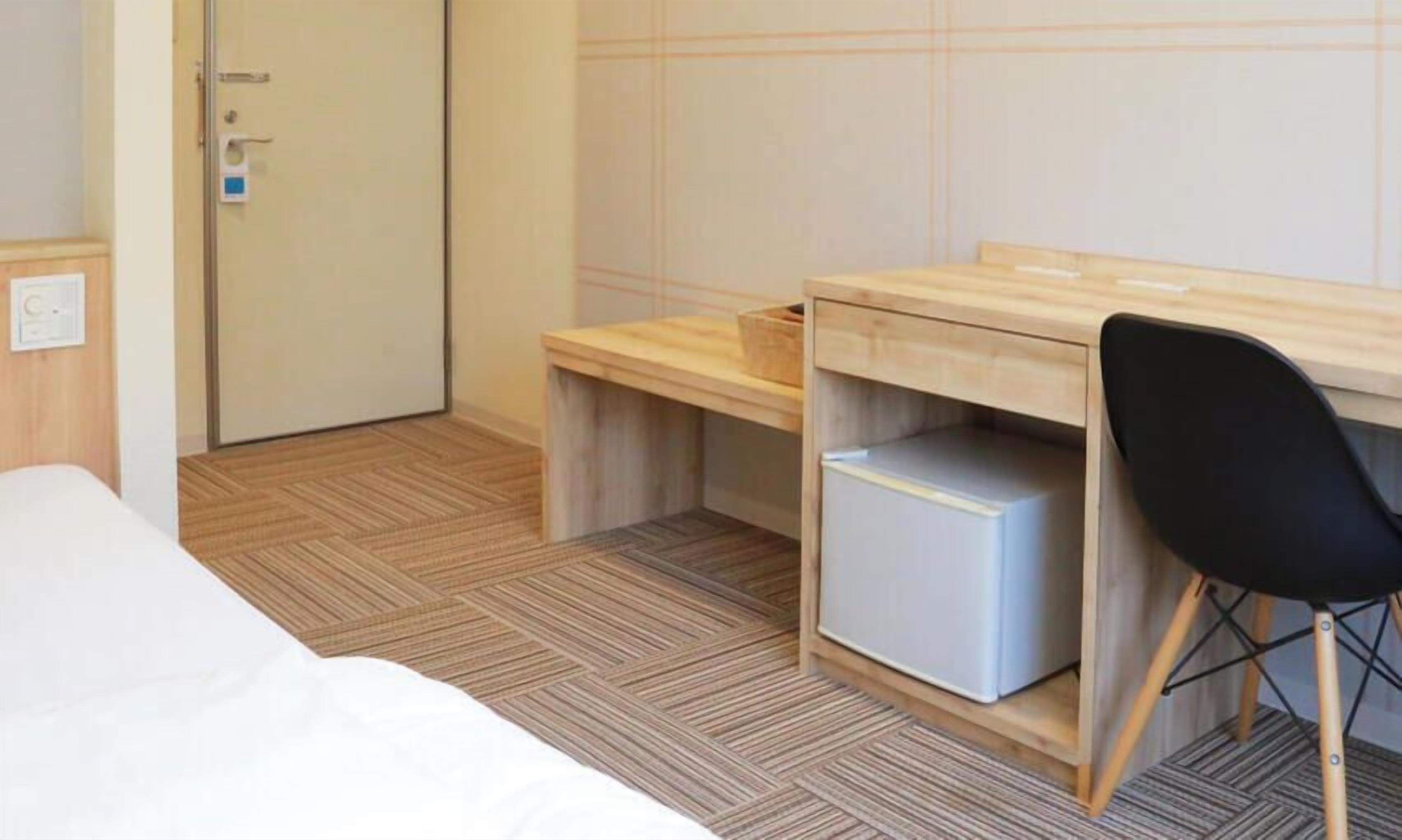 A hotel room with a bed, a wooden desk with a chair, a small white cabinet, and a door.