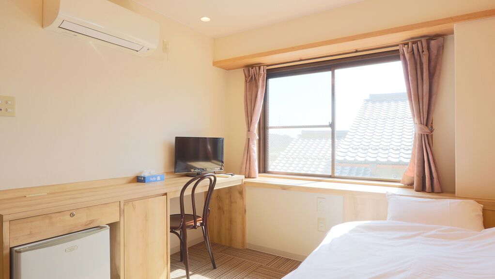 A hotel room with a bed, a desk with a computer, a chair, a small refrigerator, and a window with curtains.