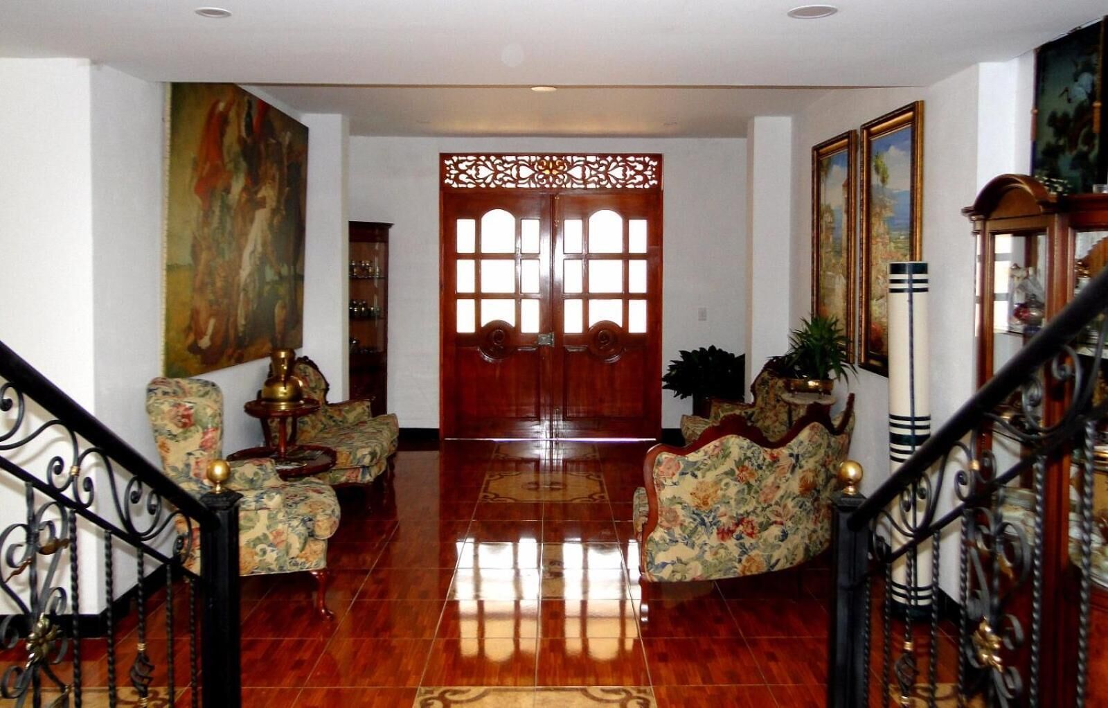 A spacious foyer with a polished wooden floor, a grand wooden door with intricate carvings, and a staircase with ornate metal railings.