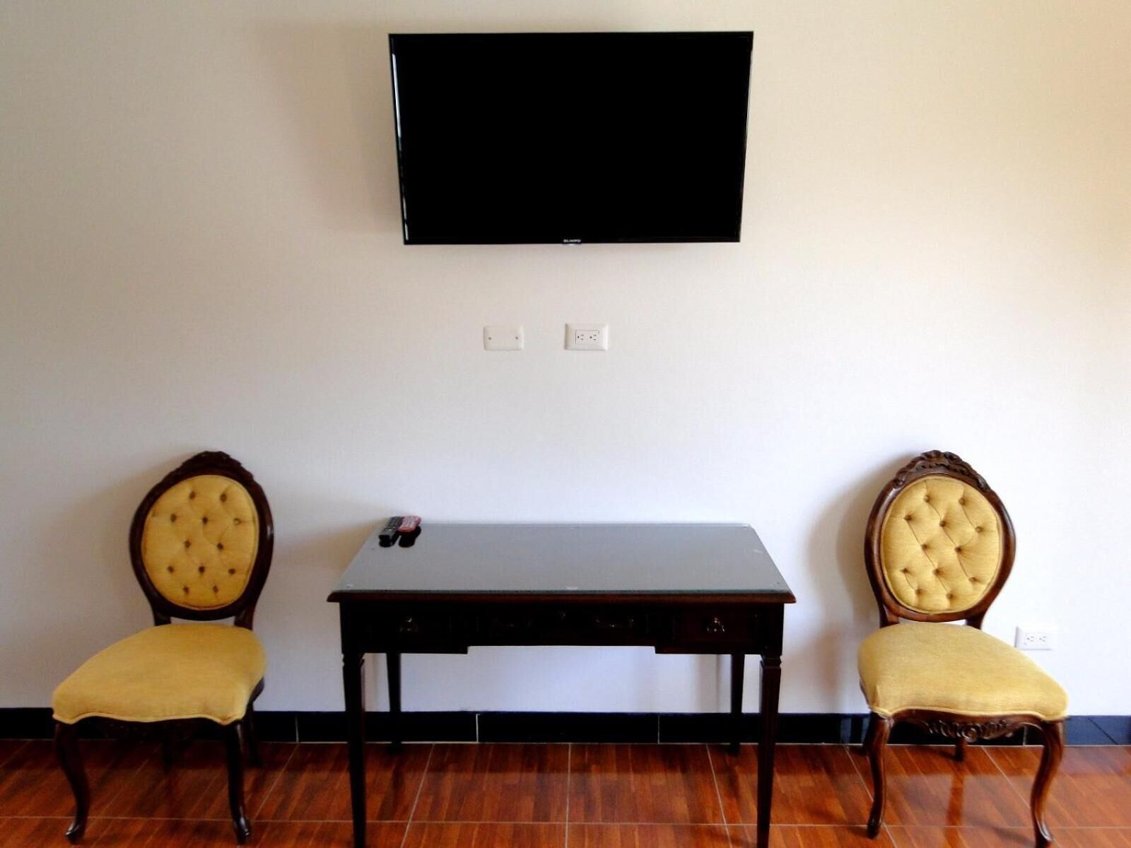 A room with a flat-screen TV mounted on the wall, a small table, and two upholstered chairs with wooden frames.