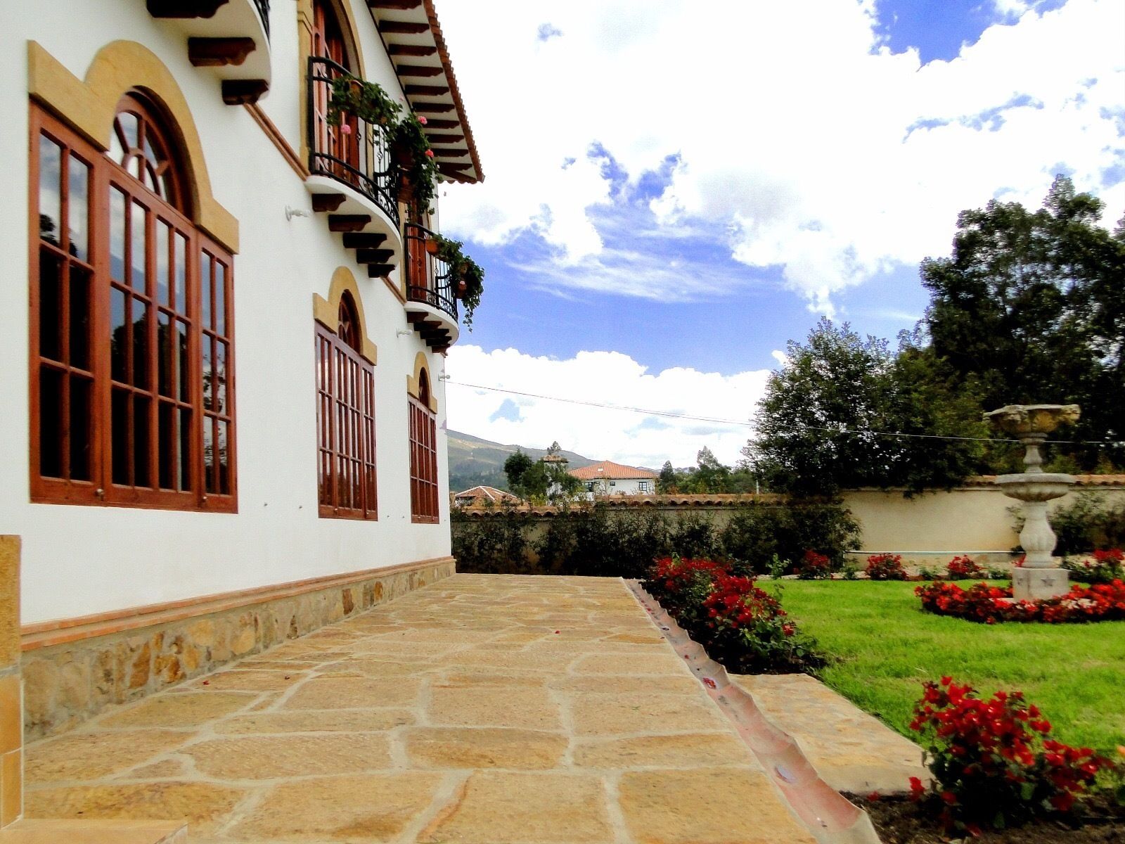 Un edificio con entrada empedrada de piedra, ventanas grandes con marcos de madera y un jardín con flores rojas y una fuente.