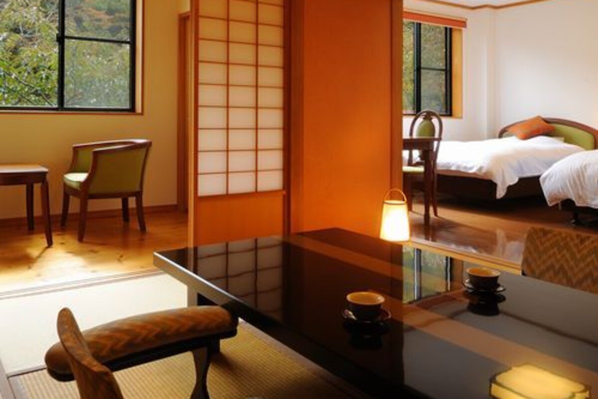 Une pièce de style japonais traditionnel, avec un sol recouvert de tatamis, une petite table basse avec un coussin, un lit avec un oreiller et une vue sur la verdure à l’extérieur.