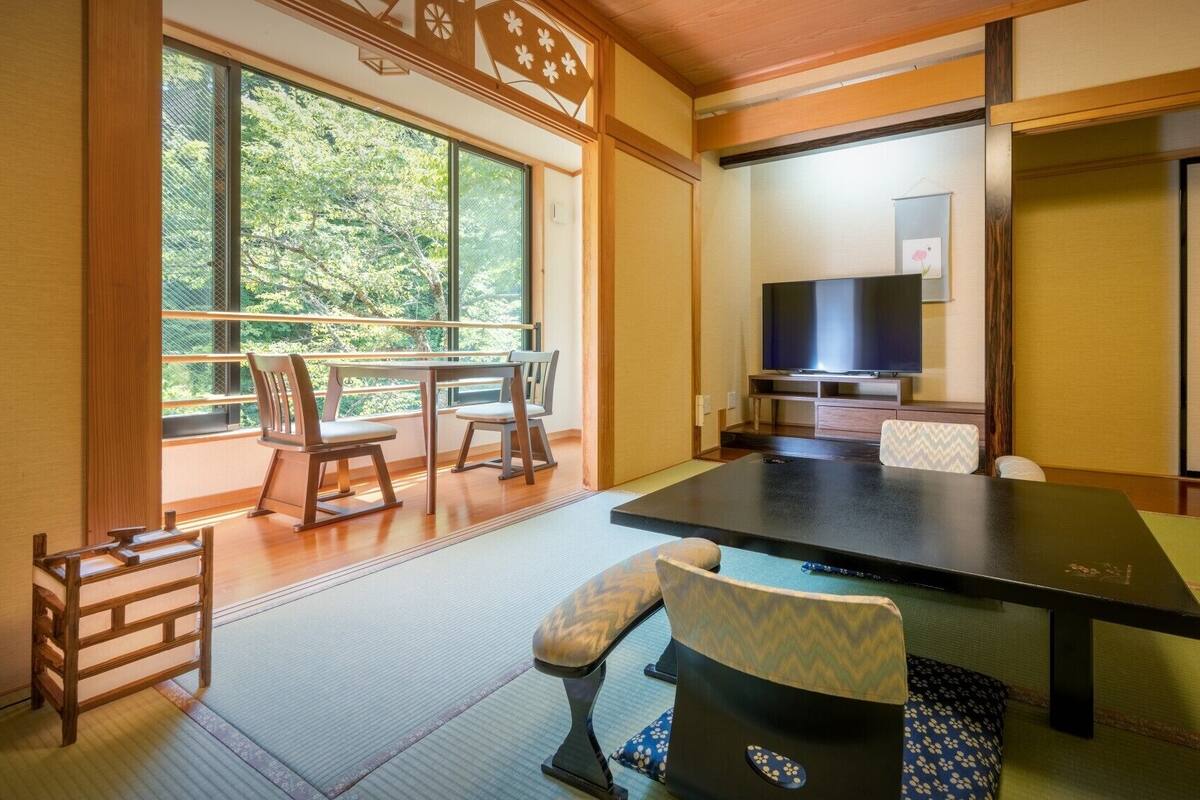 Ein traditioneller japanischer Raum mit Tatami-Bodenbelag, einem niedrigen Tisch, Stühlen, einem Fernseher und einer Schiebetür.