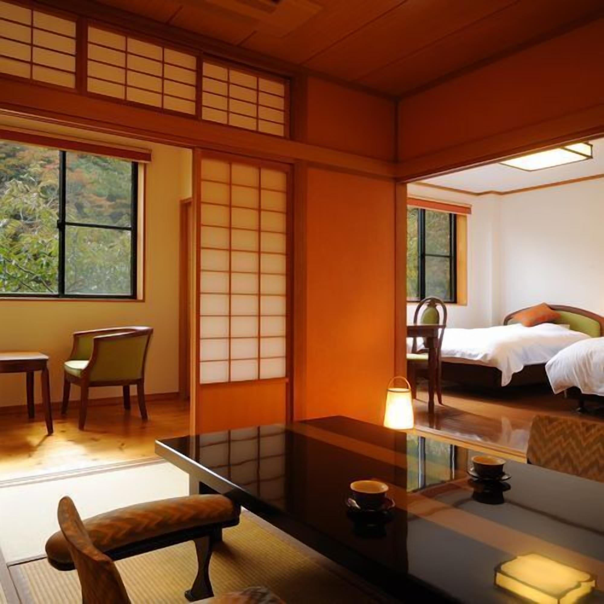 A traditional Japanese room with tatami flooring, a low table with a cushion, a bed with a pillow, and a view of greenery outside.