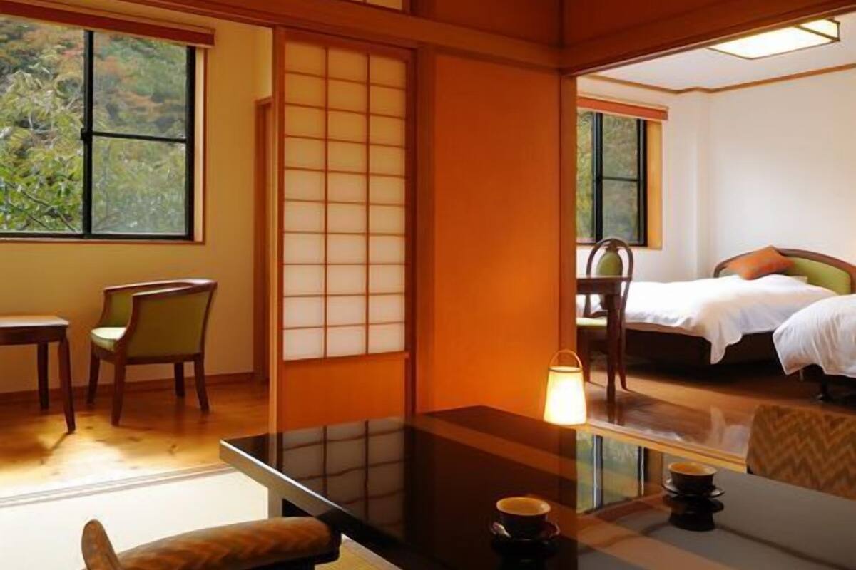 Ein traditioneller japanischer Raum mit Tatami-Boden, einem niedrigen Tisch mit Kissen, einem Bett mit Kopfkissen und Blick auf Grünfläche.
