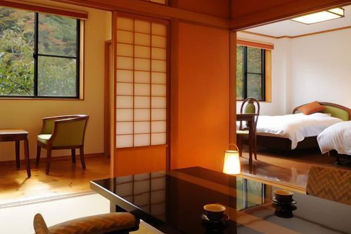 A traditional Japanese room with tatami flooring, a low table with a cushion, a bed with a pillow, and a view of greenery outside.