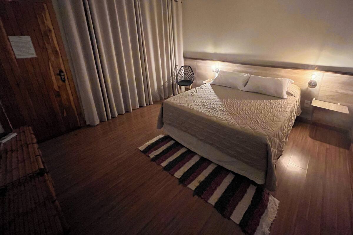 A hotel room with a bed, a chair, a nightstand, a lamp, and a window with curtains.