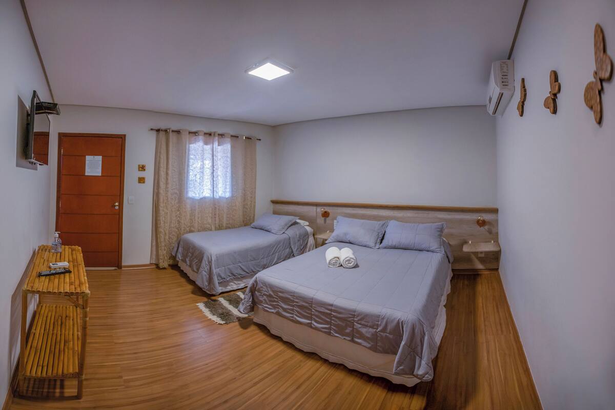 A room with two beds, a wooden table, a desk, a window with curtains, a door, and an air conditioning unit.