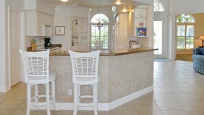 A kitchen with a bar area, white bar stools, and a view of the outside through a window. - Relaxing Waterfront Home With Pool and Steps to Beach (Marco Island)