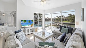 A modern living room with a large TV, comfortable seating, and a view of a pool and palm trees. - Relaxing Waterfront Home With Pool and Steps to Beach (Marco Island)