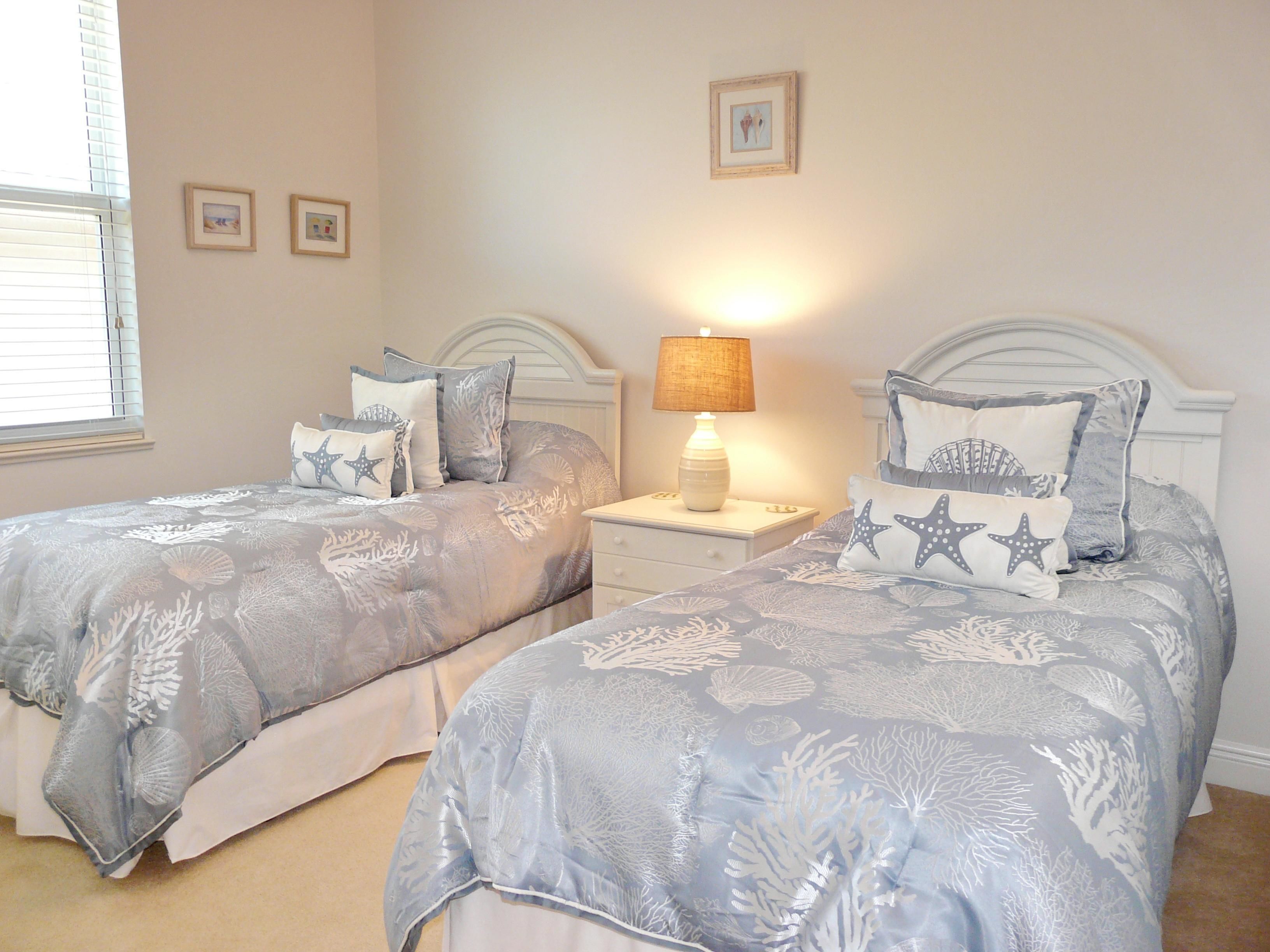 Une chambre avec deux lits, chacun agrémenté de la même literie et de coussins en forme d’étoile de mer, une table de chevet avec une lampe et des tableaux encadrés accrochés au mur.