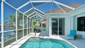 A swimming pool with a glass enclosure, a covered barbecue area, and a seating area with a view of the water. - Relaxing Waterfront Home With Pool and Steps to Beach (Marco Island)