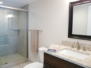 A bathroom with a glass shower enclosure, a white toilet, a sink with a marble countertop, and a mirror above the sink. - Seawinds (Marco Island)