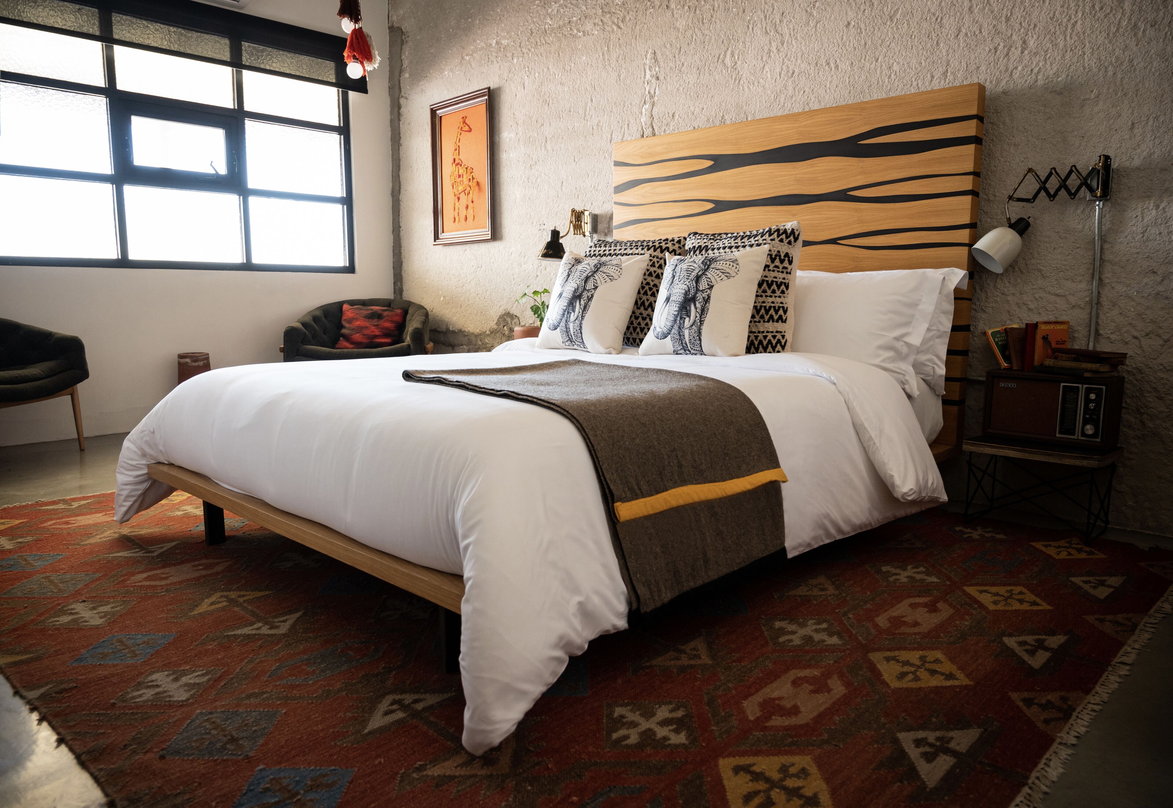 A neatly made bed with a patterned rug, a wooden headboard, and a large window.