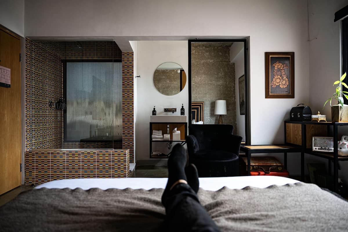 Ein Hotelzimmer mit Bett, einer Sitzecke mit Sessel, einem kleinen Tisch, einem Spiegel und Blick ins Bad mit Dusche und Waschbecken.
