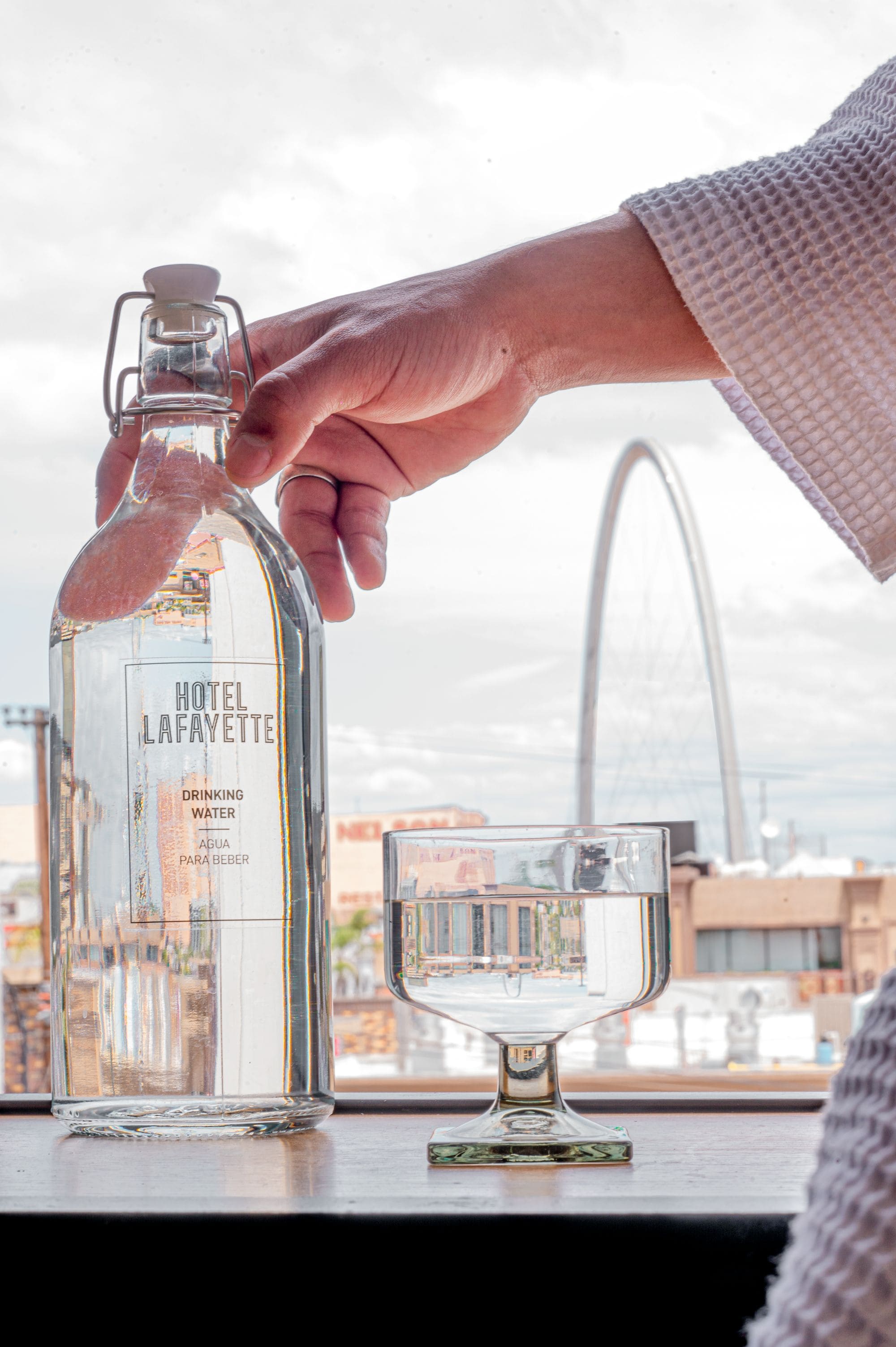 En hånd hælder vand fra en flaske med påskriften 'HOTEL LAFAYETTE' i et glas.