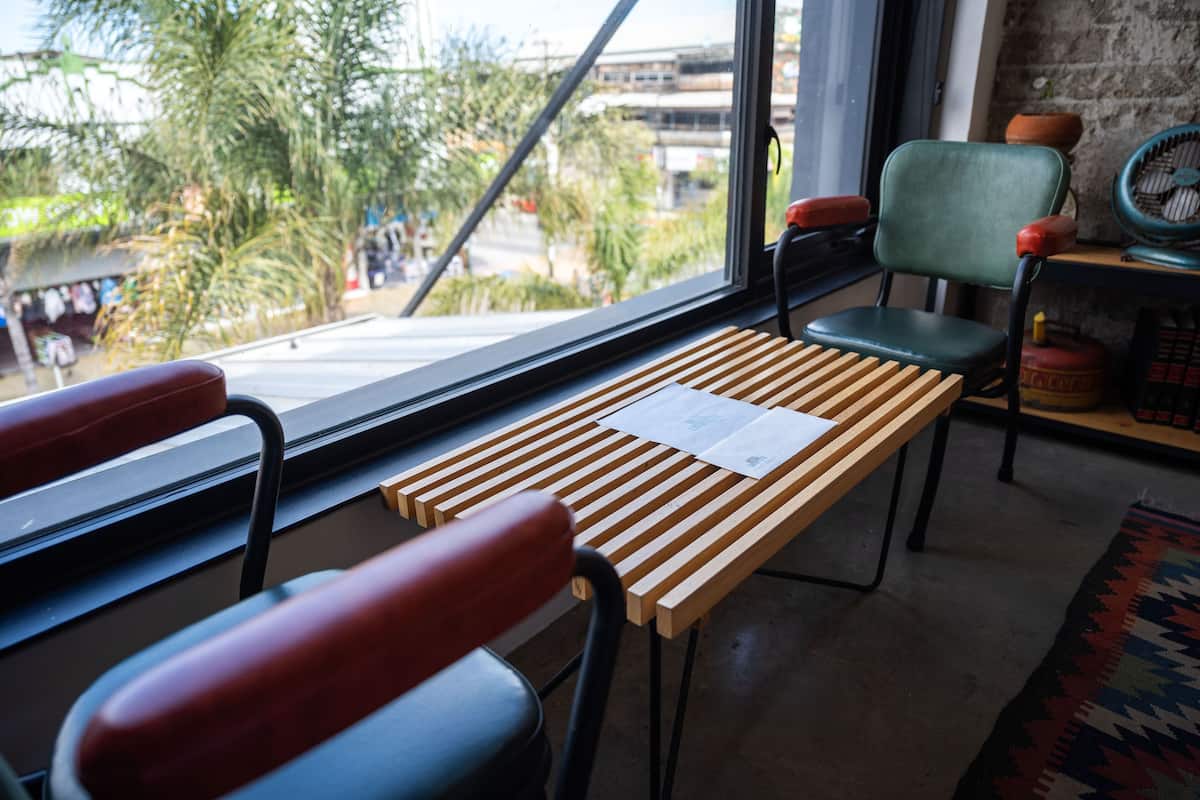 Ein Zimmer mit einem Holztisch, zwei Stühlen, einem Fenster mit Blick auf Palmen und Gebäude sowie einer Backsteinwand.