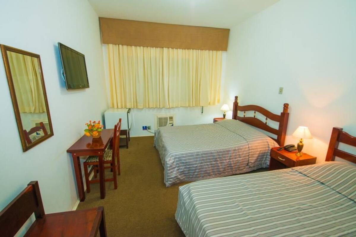 Habitación de hotel con dos camas, un escritorio, una silla, una lámpara y una ventana con cortinas.