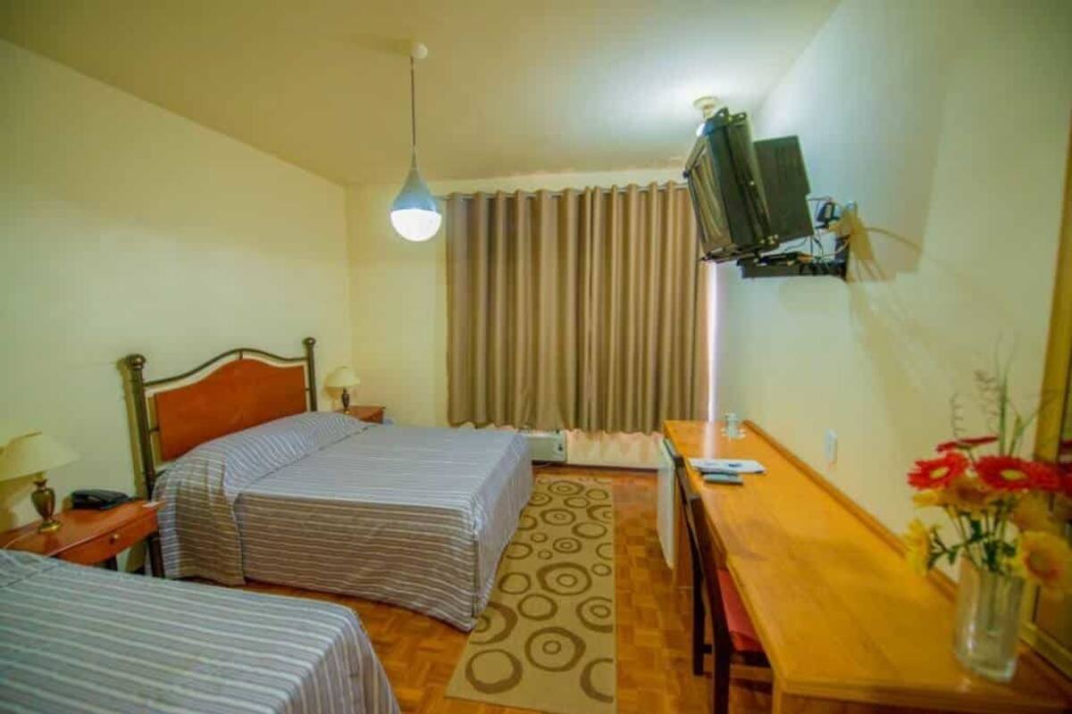 Habitación de hotel con dos camas, un escritorio de madera, televisor montado en la pared y una ventana con cortinas.