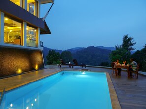 A swimming pool with a wooden deck, a dining area with tables and chairs, and a view of the mountains. - Forest Canopy (Peermade)