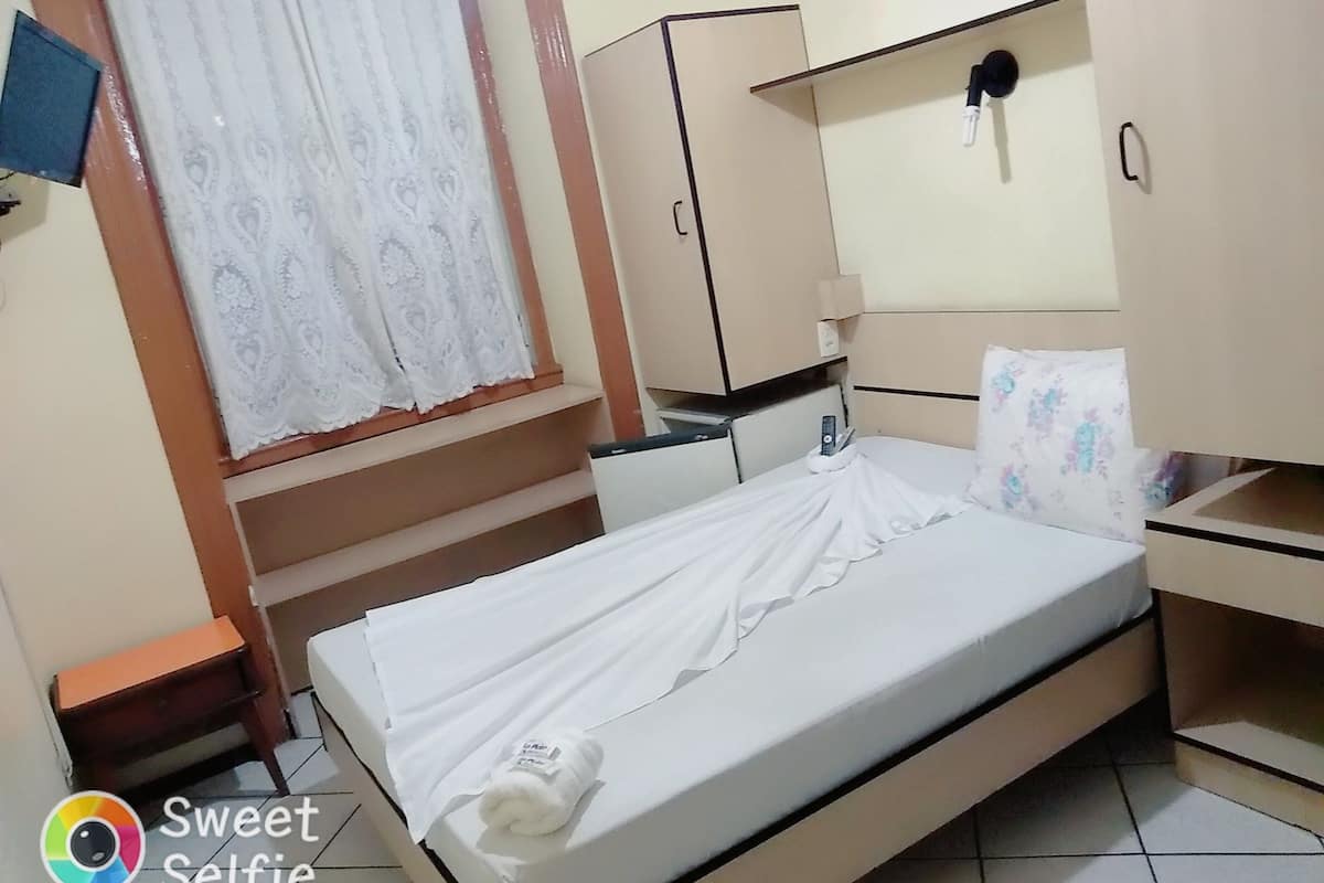 A single bed with a white mattress and a rolled towel, a small bedside table, a wardrobe, and a window with lace curtains.