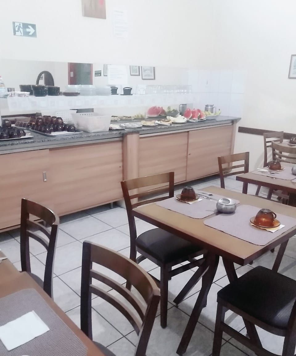 Uma área de refeições com mesas e cadeiras de madeira, uma cozinha ao fundo e uma placa indicando a saída.