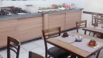 Uma área de refeições com mesas e cadeiras de madeira, uma cozinha ao fundo e uma placa indicando a saída.