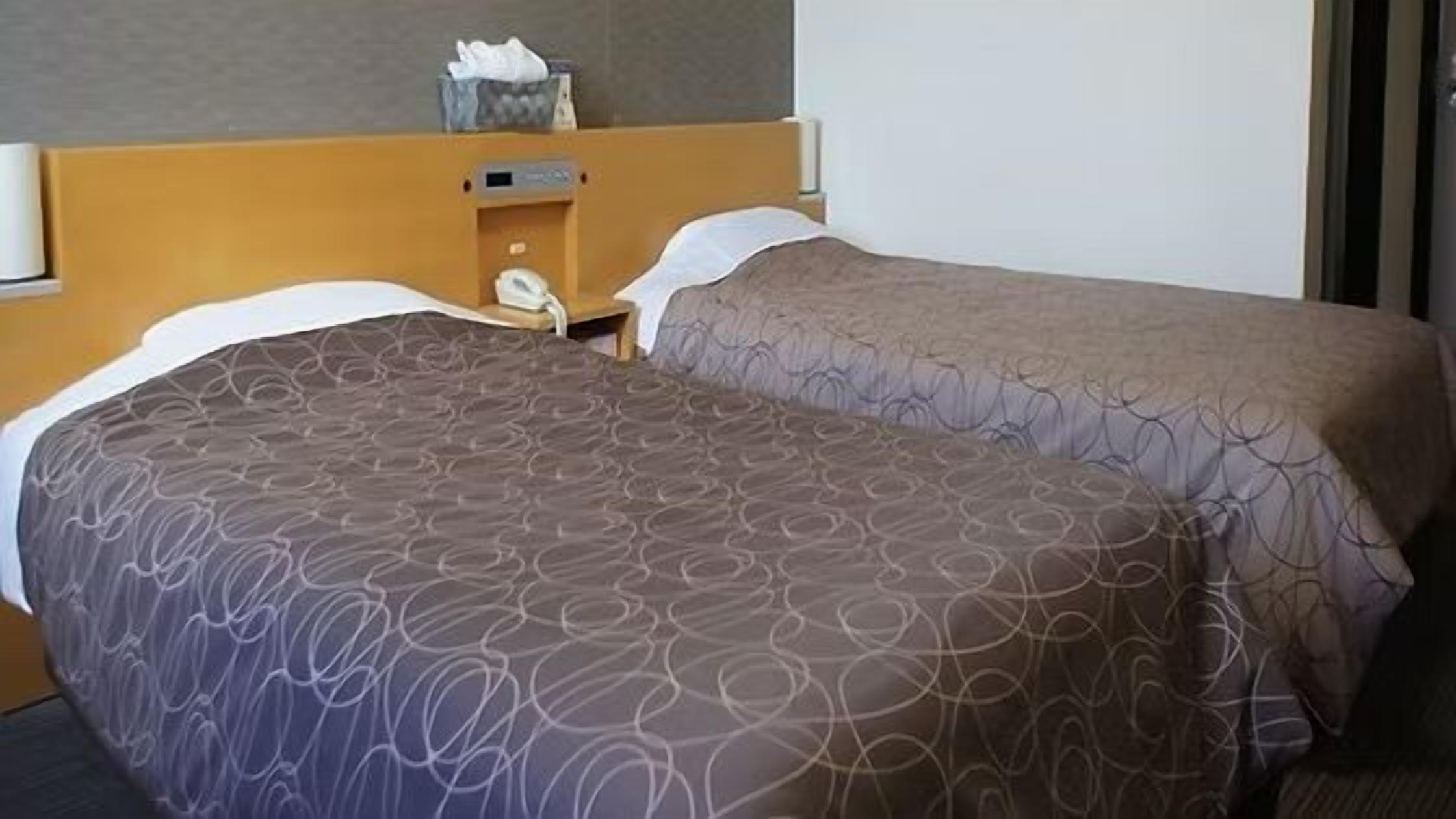 A double bed with patterned bedding, a wooden headboard, a bedside table with a lamp, and a trash bin.