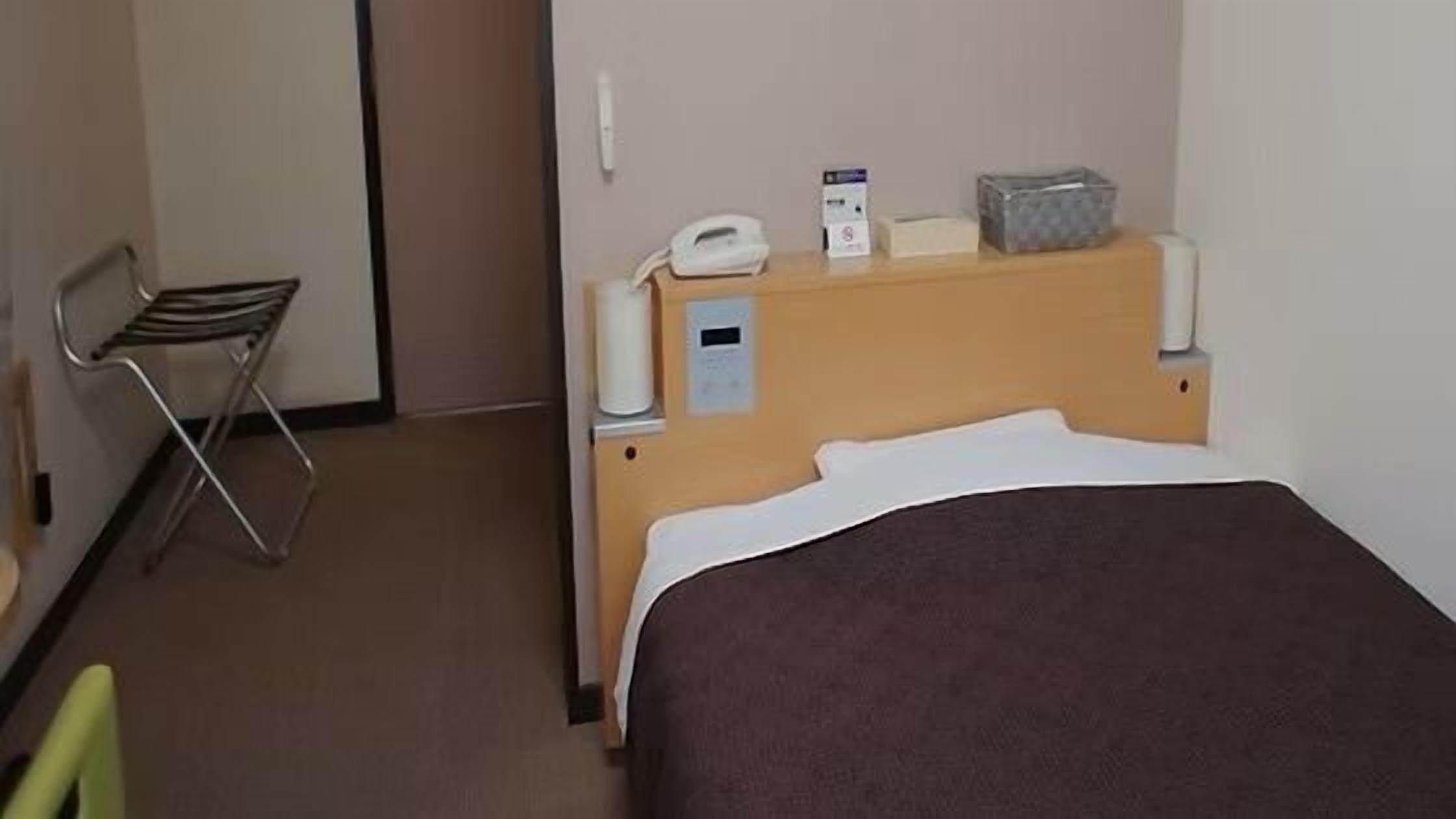 A hotel room with a bed, a chair, a bedside table with a phone and a box, and a wall-mounted thermostat.