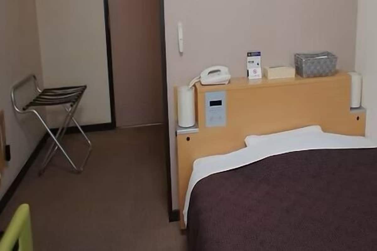 Une chambre d’hôtel comprenant un lit, une chaise, une table de chevet avec un téléphone et une boîte, ainsi qu’un thermostat fixé au mur.