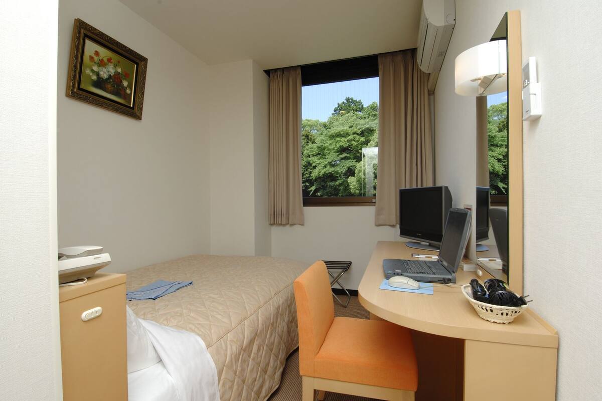 A hotel room with a bed, a desk with a computer, a chair, a desk lamp, a window with curtains, and a picture on the wall.