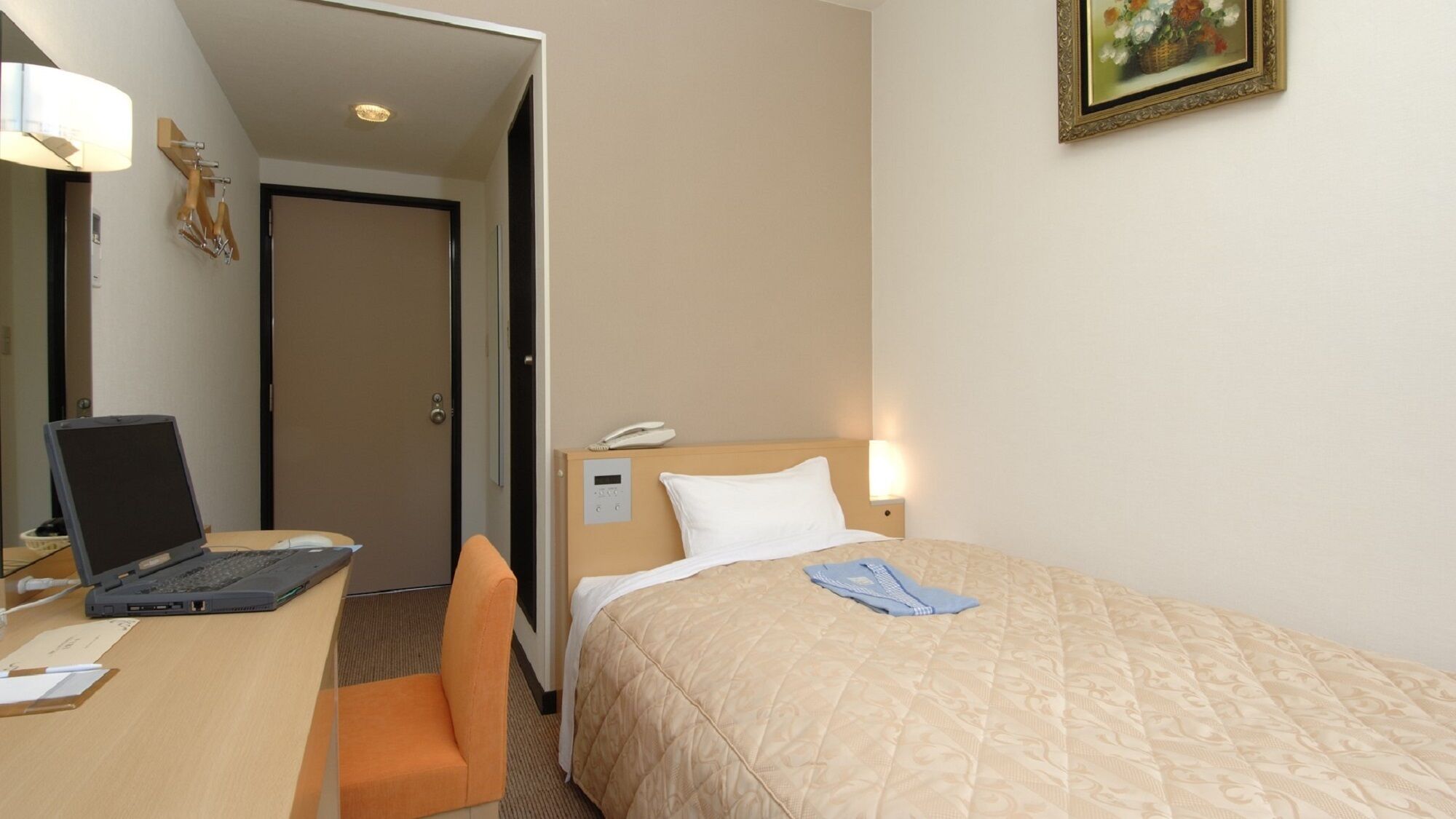 A hotel room with a bed, a desk with a laptop, a chair, a bedside table with a lamp, and a painting on the wall.