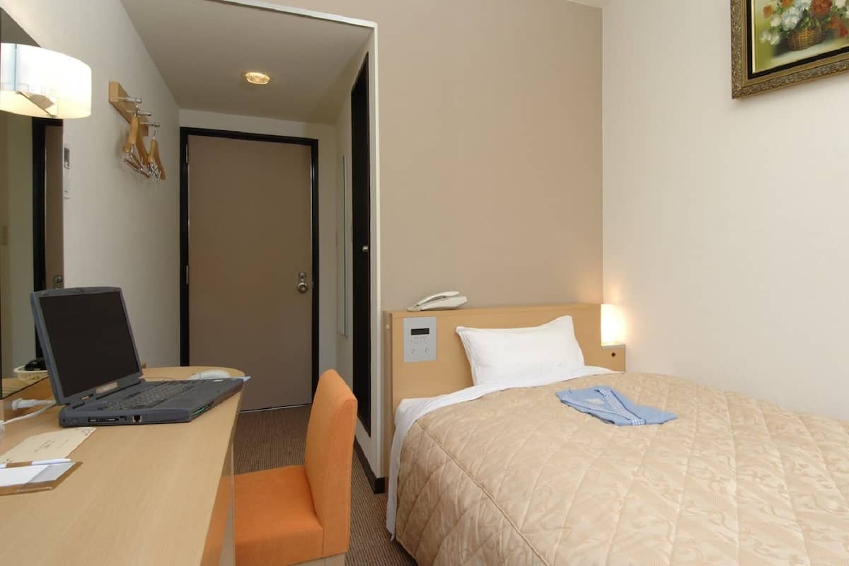 A hotel room with a bed, a desk with a laptop, a chair, a bedside table with a lamp, and a painting on the wall.