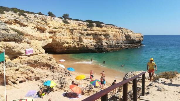 Ein Strand mit felsigen Klippen, klarem türkisblauem Wasser und badenden Personen.