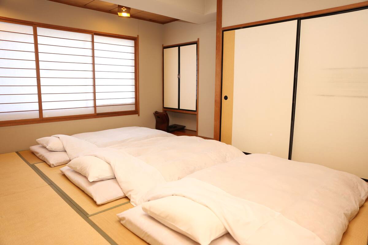 Um quarto japonês tradicional com piso de tatame, uma cama grande com roupa de cama branca, uma porta de correr e uma janela com biombos shoji.